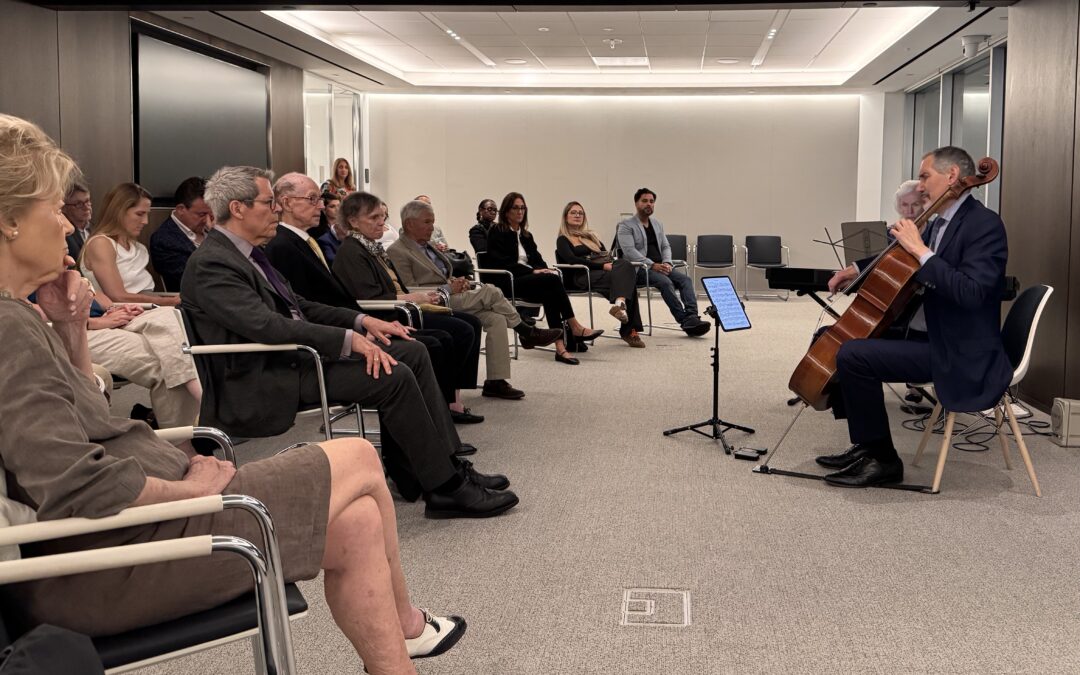 A modern room with an audience of thirty adults seated in a semi-circle facing a man playing a cello and a woman playing the piano.